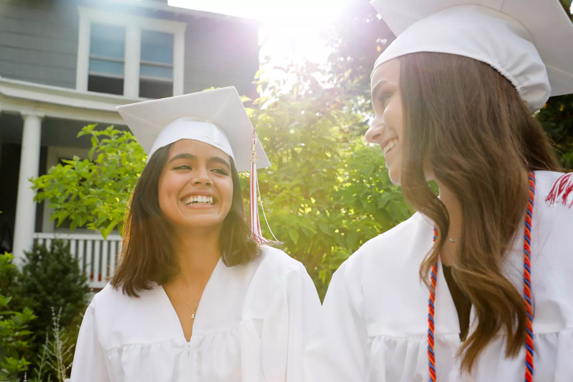two smiling graduates in white graduation gowns outside house